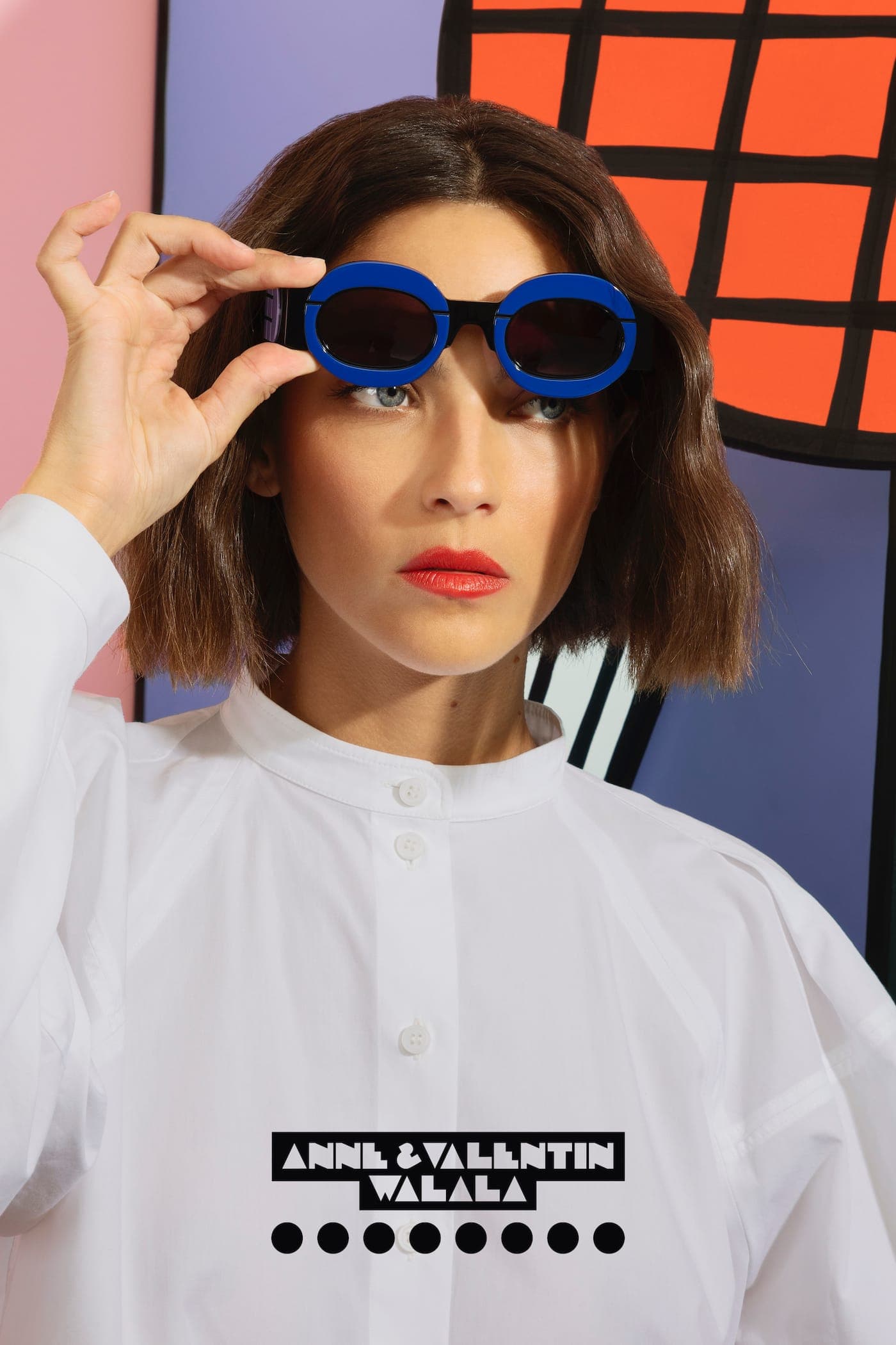A woman holding up bold blue round Anne & Valentin sunglasses from the Walala collection, against a colourful graphic backdrop