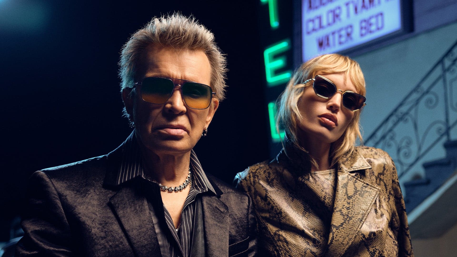 A man and a woman wearing Cutler and Gross sunglasses in front of a neon hotel sign, night scene